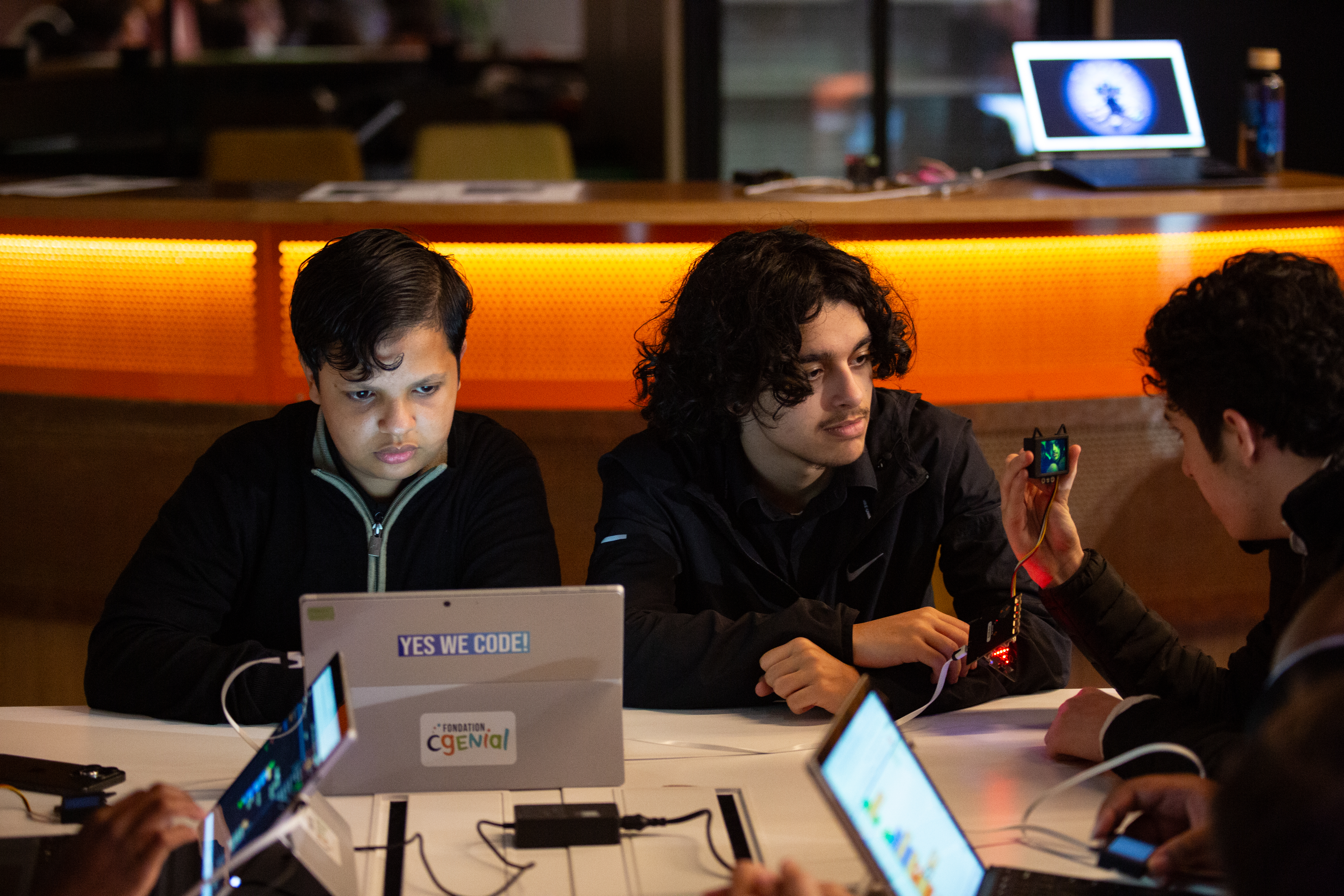 Des étudiants travaillent en groupe autour d'une table avec des ordinateurs portables. L'un d'eux manipule un petit composant électronique. Un autocollant 'YES WE CODE!' est visible sur un ordinateur.