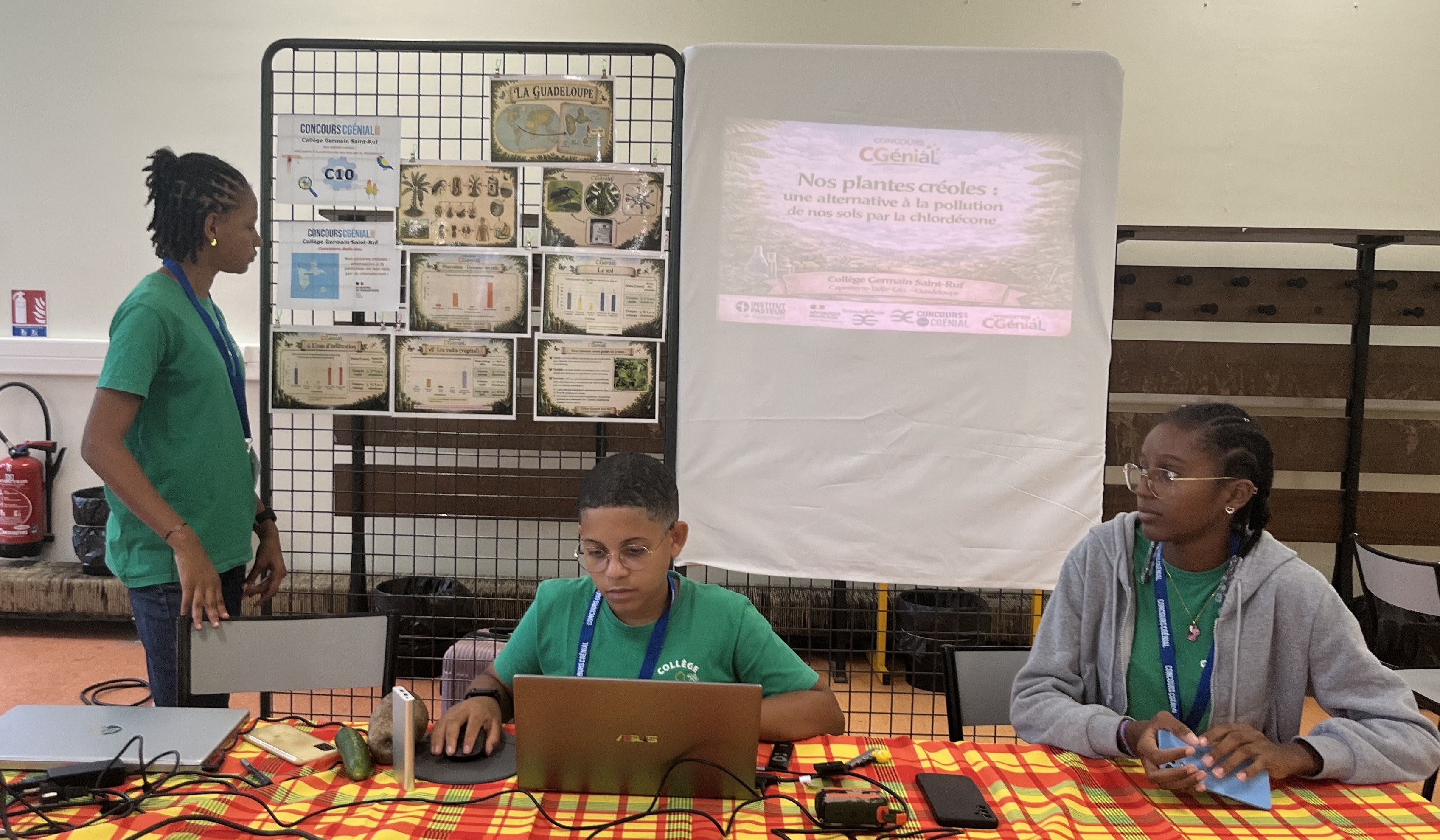 Trois collégiens présentent un projet scientifique lors du concours CGénial. Ils se tiennent autour d'une table recouverte d'une nappe madras jaune et rouge, sur laquelle sont disposés un ordinateur portable, des échantillons de plantes et des flacons. En arrière-plan, un panneau d'affichage et un écran de projection affichent le titre 'Nos plantes créoles : une alternative à la pollution de nos sols par la chlordecone'."