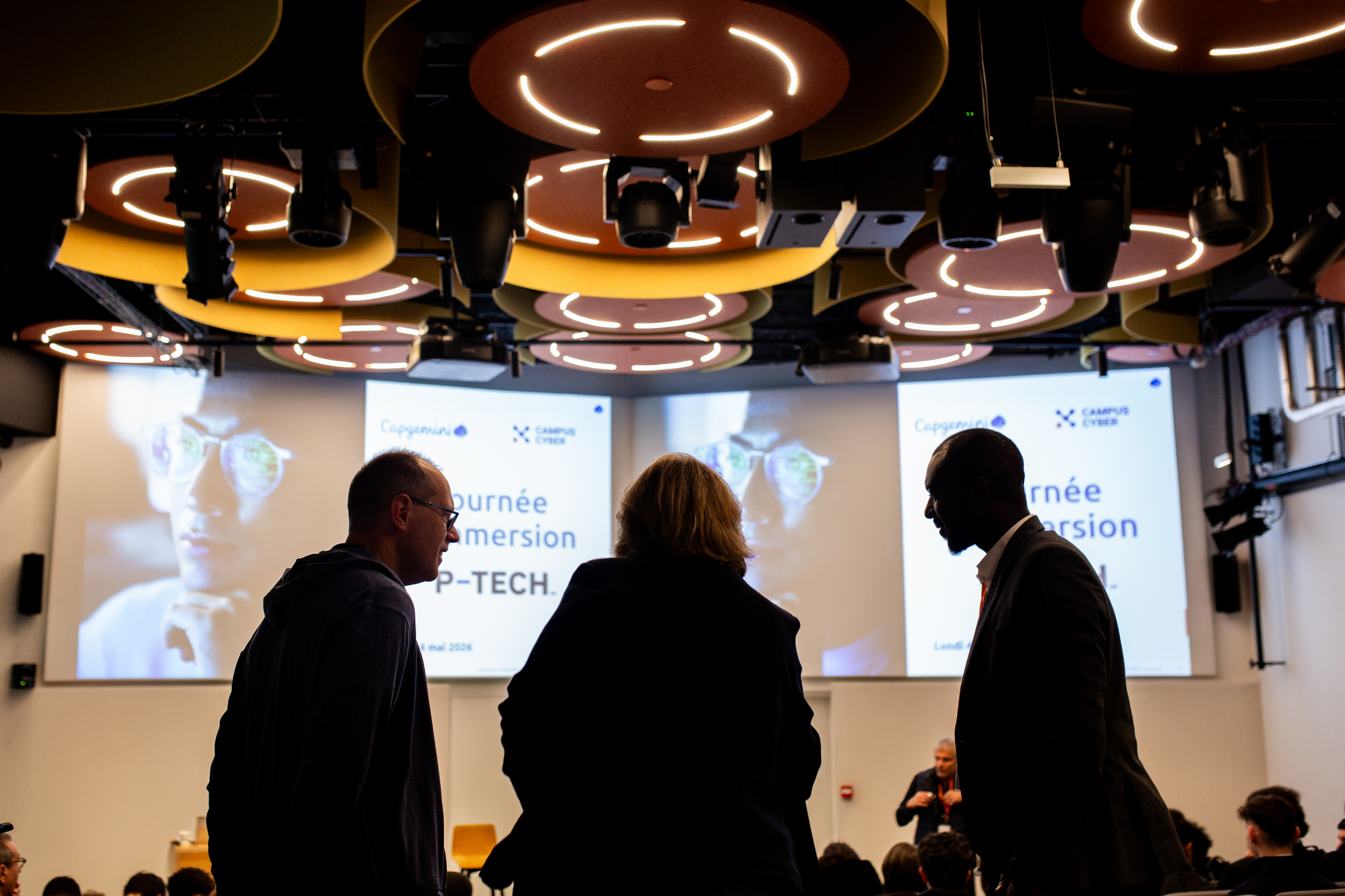 Event P-Tech & Capgemini - mai 2026 Trois personnes discutent en silhouette au premier plan d'une salle de conférence. En arrière-plan, un grand écran affiche 'Journée d'Immersion P-TECH' sous les logos de Capgemini et du Campus Cyber.